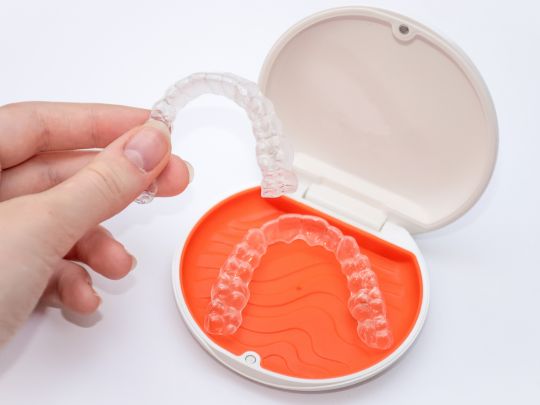 A person's hand holds a clear, plastic dental aligner above an open case with an orange interior. Another aligner rests inside the case.