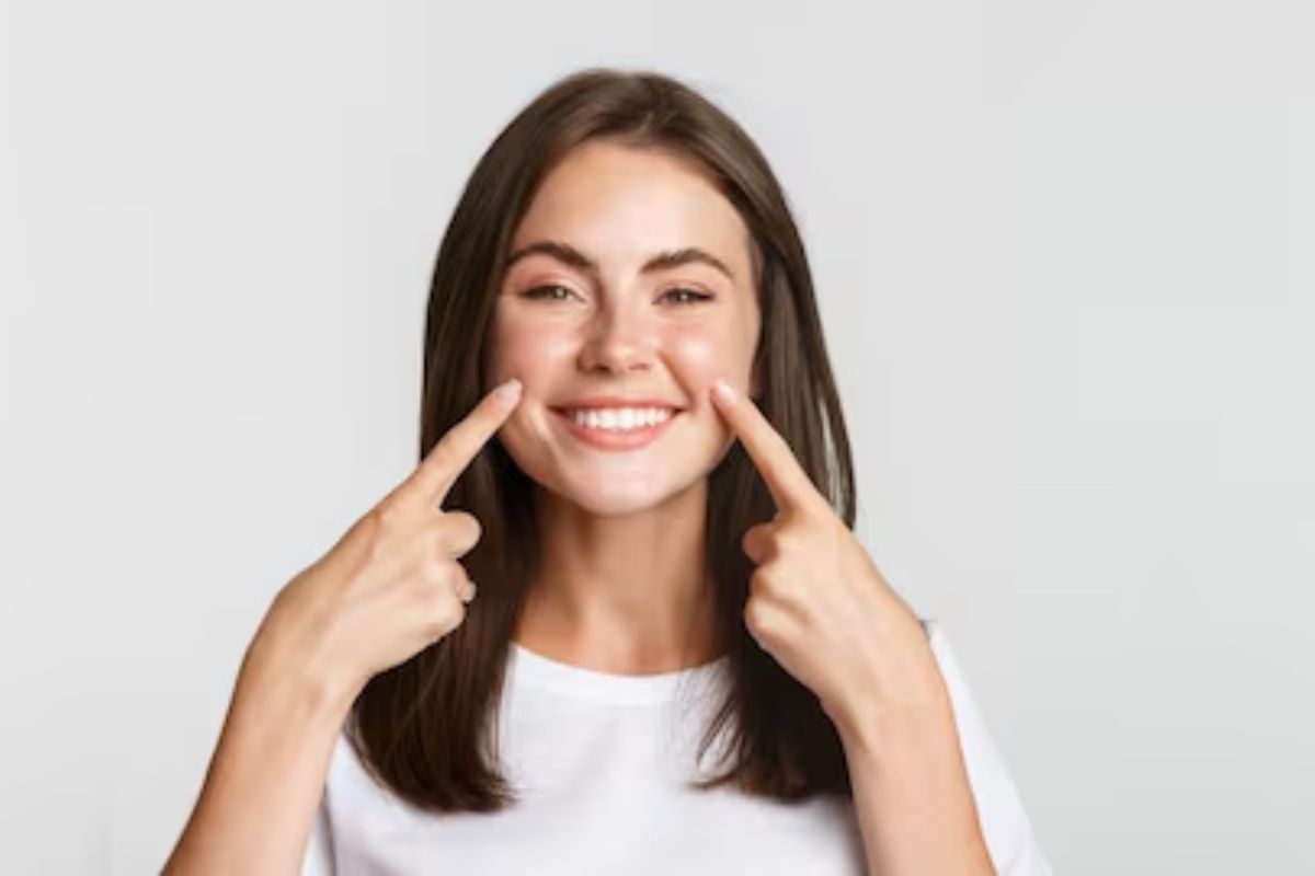 Teeth Cleaning Tulsa Young woman smiling brightly while pointing to her teeth against white background.