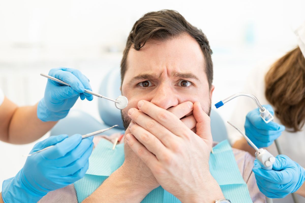 Anxious male patient covering mouth during dental examination with dentist tools and suction.